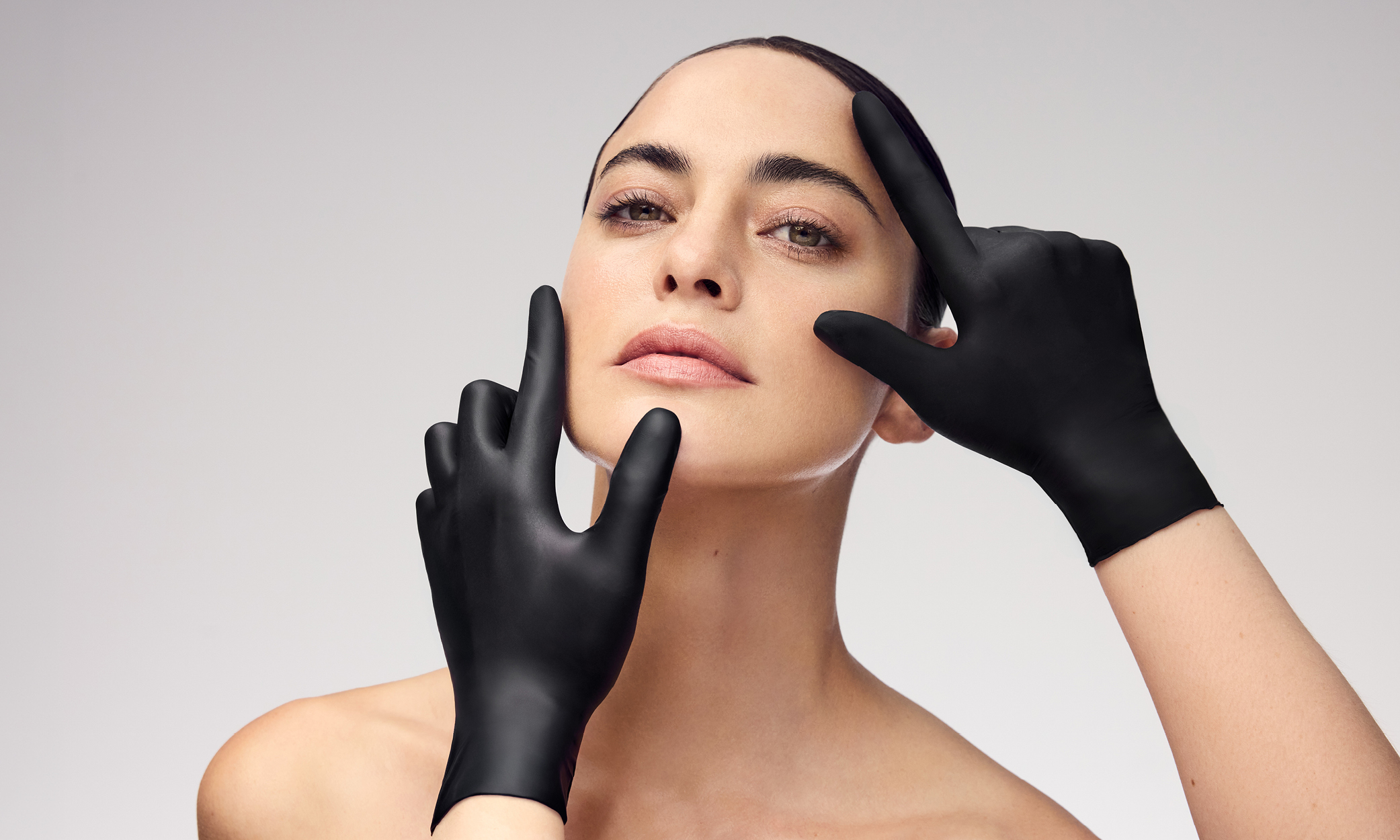 Gesicht einer Frau mit ebenmäßiger Haut, sanft umrahmt von zwei schwarz behandschuhten Händen – professionelle Kosmetik.