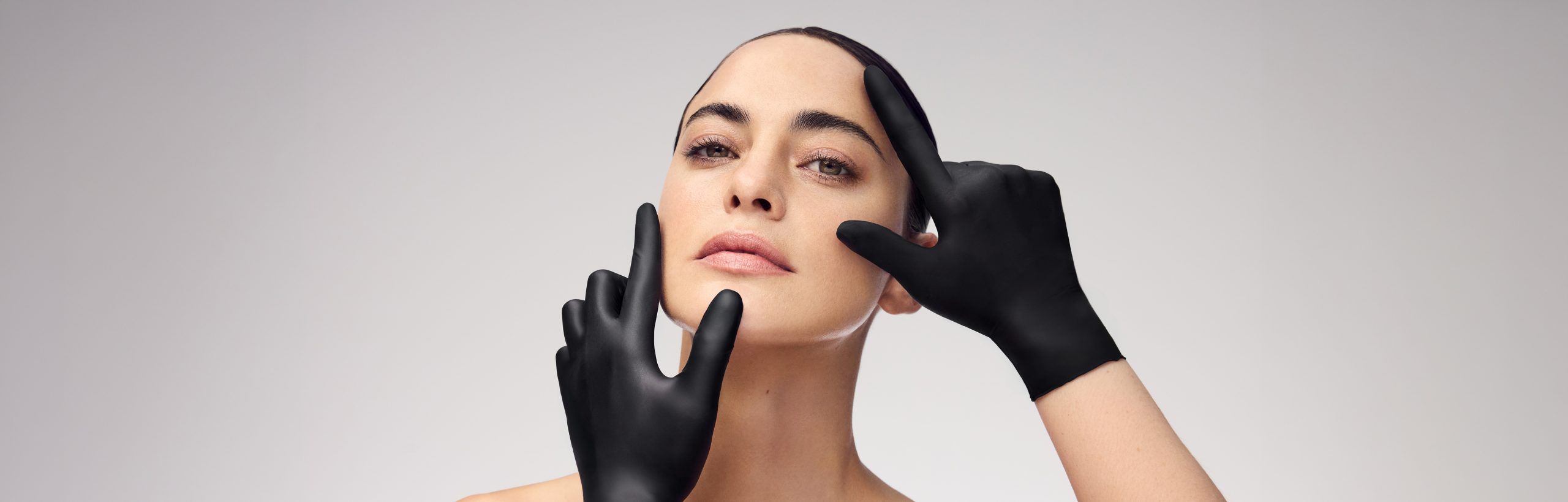 Gesicht einer Frau mit ebenmäßiger Haut, sanft umrahmt von zwei schwarz behandschuhten Händen – professionelle Kosmetik.
